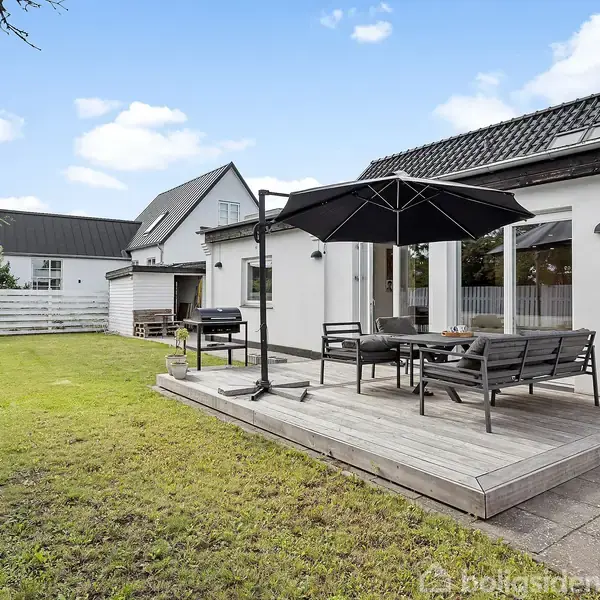 Terrasseområde med parasol og havemøbler står på en træplatform i en græsplæneomgivet have ved et hvidt hus. Der er grill ved siden af terrassen, omgivet af hegn og træer.