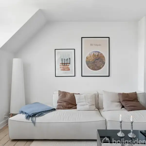 En hvid sofa med puder står i stuen. To vægplakater hænger over sofaen med teksten "Bali Nights". Rummet er lyst med grønne planter og trægulv.
