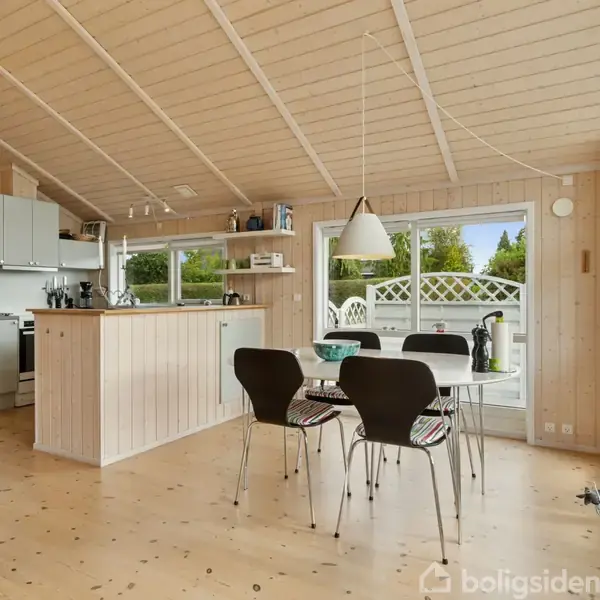 Køkken-alrum med spisebord og fire stole, åbent til et hvidt køkken med komfur og køleskab, omgivet af lyse trævægge og store vinduer, der giver udsigt til en terrasse.