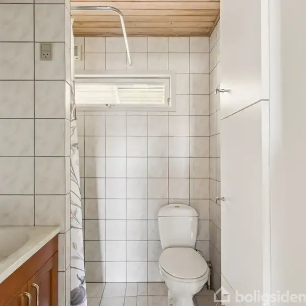 Toilet skål placeres ved siden af vask med træskab. Bruser og fliser fylder væggenes overflade. Lystræ-loft skaber et lyst badeværelse.