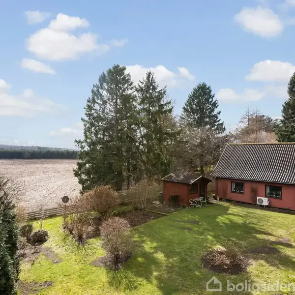 Rødt hus står stille i en have med grønne planter og buske; omgivet af høje træer og græssende marker under en blå himmel med lette skyer.