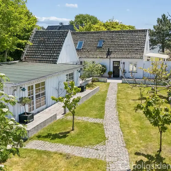 Et hvidt hus med skråt tag står i en have. Huset har vinduer og en gangsti fører til en terrasse. Haven er omkranset af små træer og grønt græs.