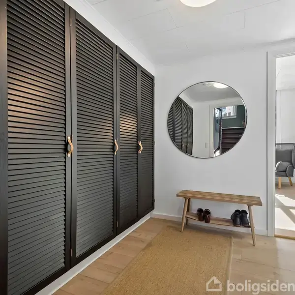 Sort skab med louvered døre står langs en væg. Rundt spejl og træbænk med sko nedenunder. Tæppe på trægulv. Åben dør leder til lysere rum.