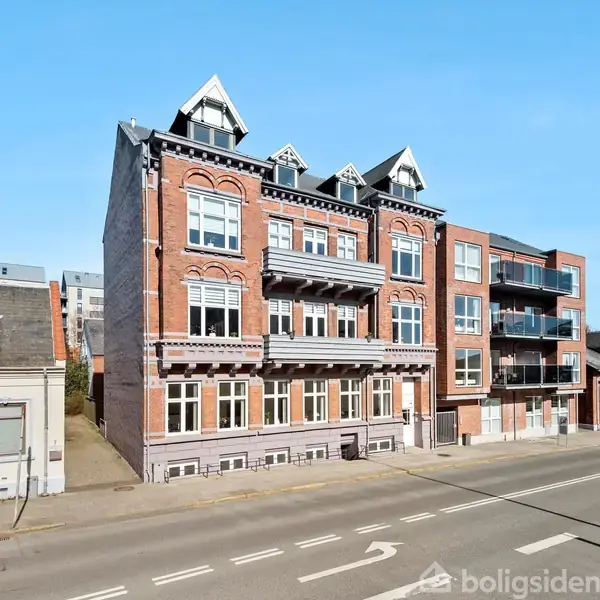 Bygning med murstensfacade står stille langs en bred vej i solskinsvejr. Omkringliggende bygninger viser et bymiljø, med flere etager og klassisk arkitektur.