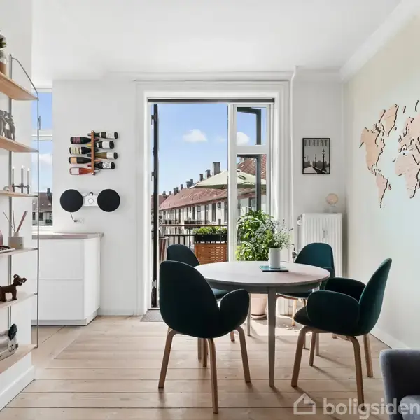 Et moderne spiseområde med et rundt bord og fire stole i et lyst rum. Ved siden af er en træhylde med dekorationer. En åben dør fører til en balkon med udsigt over bygninger. En verdenskort vægdekoration hænger på væggen.