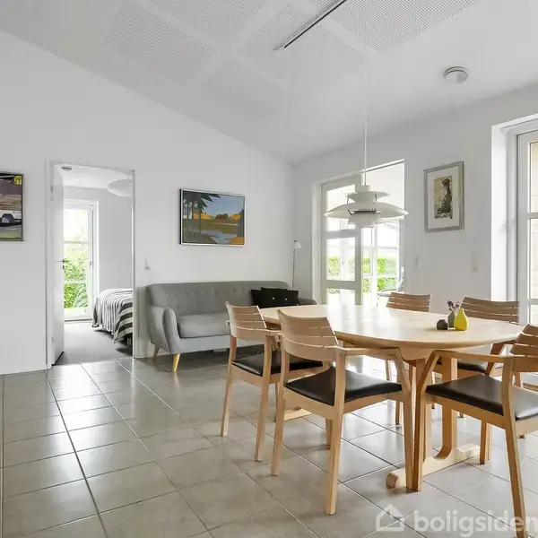 Et lyst spiseområde med et rundt træbord og seks stole. Bagved står en sofa. Rummet har store vinduer og vægkunst. En dør åbner til et soveværelse med en seng.