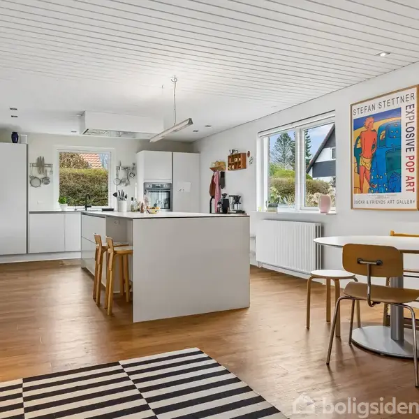 Et moderne køkken med en stor ø i midten omgivet af stole. Et bord med stole står ved siden af. Væggene har plakater og et vægur. Store vinduer giver udsigt til haven.