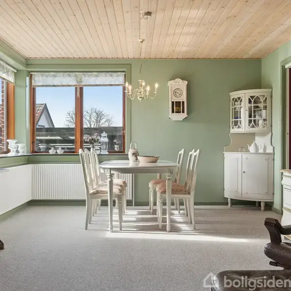 Stue med træloft og grønne vægge. Flere stole er placeret omkring et bord i hjørnet. Et stort, udskåret skab står ved et vindue med udsigt til have.