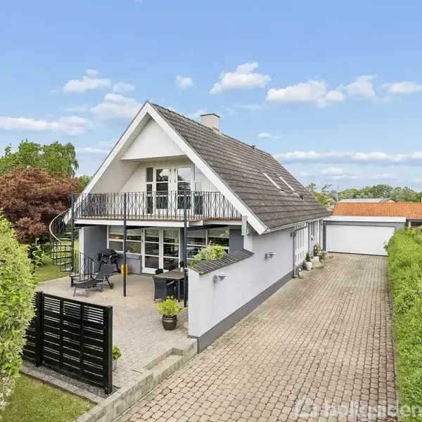 Et moderne køkken med hvide skabe og mørke fliser. Fokus er på en stor køkkenbordplade med planter ved vinduet. To ophængte skærme er monteret på væggen.