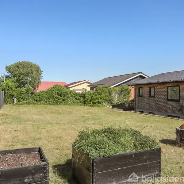 Træhøjbede står på en græsplæne i en baghave ved siden af et murstenshus. Der er en lav murstensovn i nærheden og et hegn til venstre.