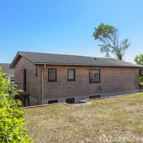 En muret etplansvilla står i rolig haveomgivelser med græsplæne og buske. Solrig himmel og træer ses i baggrunden.