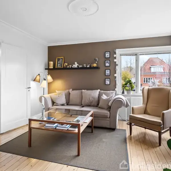 Sofa med puder står i hyggelig stue, omgivet af lamper, billeder på væggen og grønne planter. Træbord foran sofa, store vinduer slipper lys ind; parketgulv dækker gulvet.