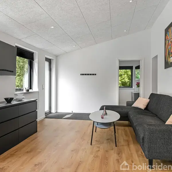 En moderne stue med en grå sofa, et lille rundt sofabord, og en sort skænk. Væggene er hvide med en farverig løveplakat. Vinduer tilbyder naturligt lys.
