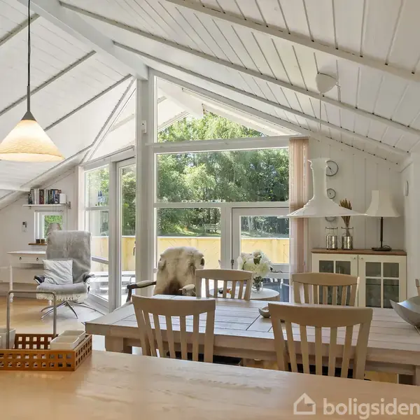 Stue indrettet med spisebord og stole i træ, stor glasvindue slipper naturligt lys ind, udsigt til terrasse og grønne træer udenfor. Pejs og moderne lamper tilføjer hygge.