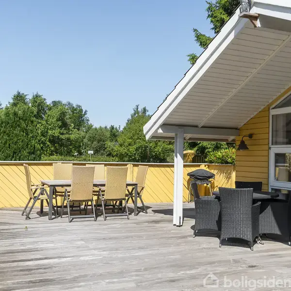 Træterrasse med spisebord og seks stole står under åben himmel. En grill er placeret i hjørnet. Omgivet af grønt løv og en gul husmur med store vinduer.