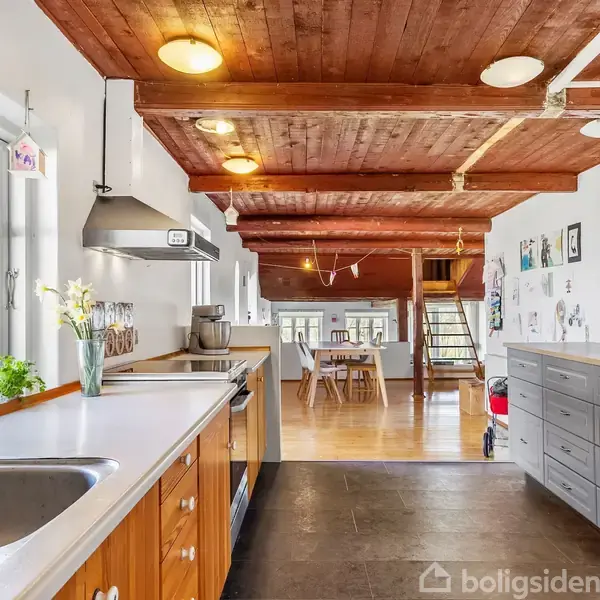 Køkken med træloft og grå fliser, hvor køkkenbordet har blomster og diverse køkkenudstyr. I baggrunden ses en spisebord med stole i et rummeligt hjem med store vinduer og lyse træmøbler.