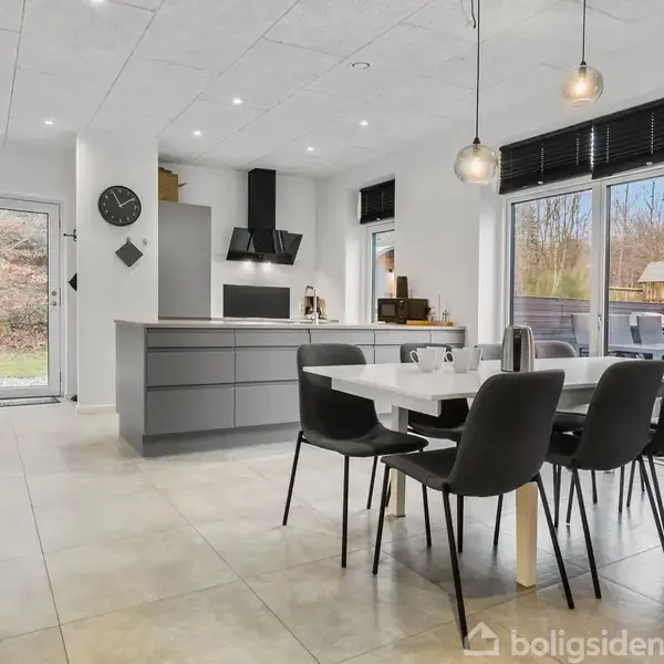 Et moderne køkken-alrum med grå køkkenø, sort emhætte, og spisebord med seks sorte stole. Store vinduer giver udsigt til en terrasse og natur udenfor. Vægur hænger til venstre.