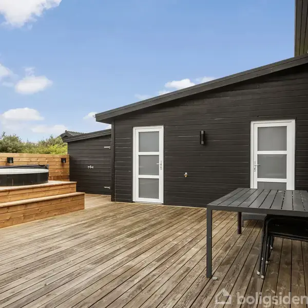 Sort træhus med to hvide døre, en træterrasse og et bord med stole. En indbygget jacuzzi i højre side omgivet af træ, under en blå himmel med få skyer.