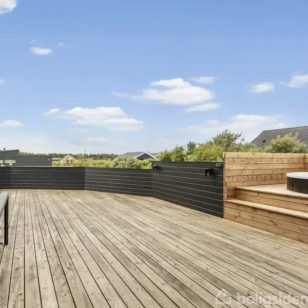 Stor terrasse med trædæk, bord med stole, og en indbygget træbænk rundt om et spabad. Omkring ses flere huse og grønne buske under en blå himmel med få skyer.