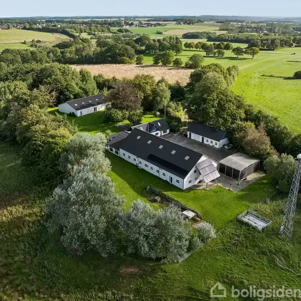 En gård, omgivet af træer og marker, findes centralt i billedet. Bygningerne har mørke tage, og der er en mast i højre side. Landskabet er grønt og bakket.