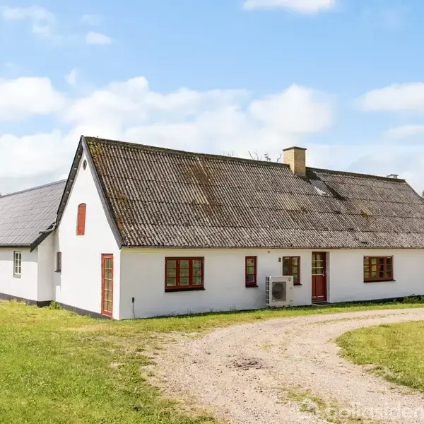 Hvidt hus med røde karme står midt i en frodig have. Grusvej leder til hoveddøren, omgivet af træer og grønne områder under en blå himmel.