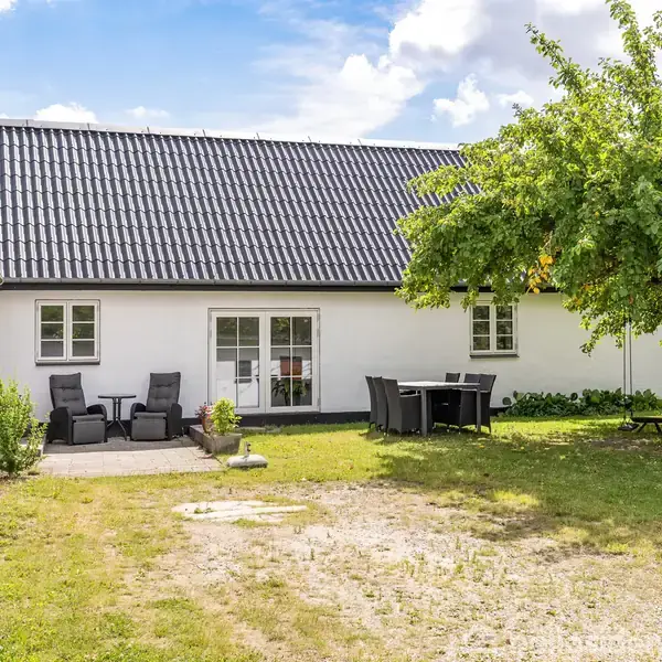 Hvidt hus med sort tag står i en have. To stole og et bord er placeret på en terrasse, mens et træ giver skygge ved et andet bord.
