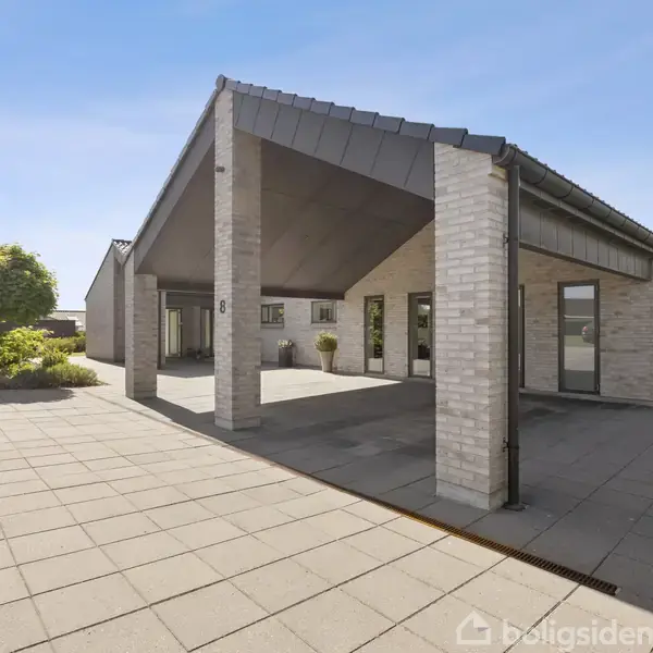Moderne hus i hvid mursten med skrånende tag, står i sollys på en stor flisebelagt terrasse. Grønne buske omkranser stedet i forstadsomgivelser. Nummer “8” ses på en mur.