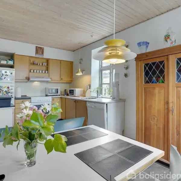 En køkken med spisebord. Der ligger sorte dækkeservietter og en blomstervase på bordet. Baggrunden indeholder skabe, køleskab og køkkenudstyr. En lampe hænger over bordet.