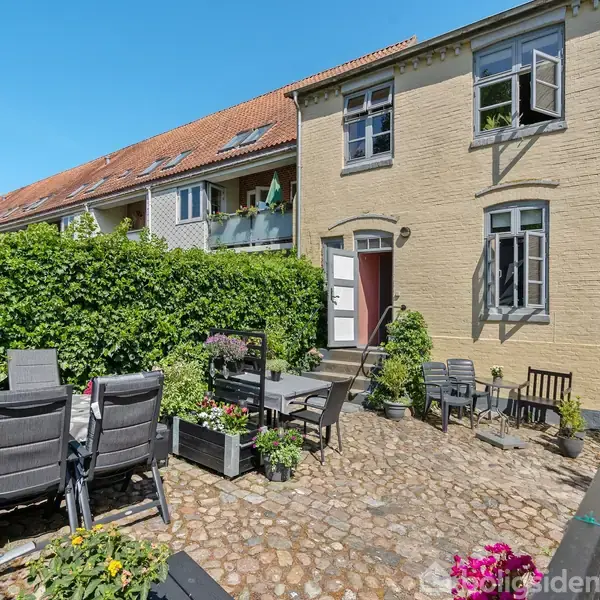 En solrig gårdhave indeholder udendørsmøbler og blomsterkrukker. Omkranset af en grusbelagt terrasse, ses en to-etagers bygning med åbne vinduer i baggrunden. Der er en høj hæk ved siden af bygningen.