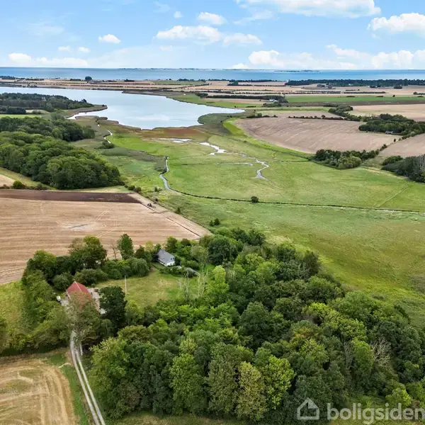 Floder og skove strækker sig gennem et åbent landskab med marker og en stor sø i baggrunden under en blå himmel med hvide skyer.