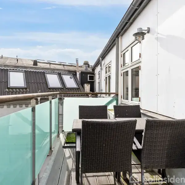 En balkon med glasrækværk har et bord og fem flettede stole. I baggrunden ses et hvidt bygningskompleks og flere tagvinduer under en klar blå himmel.