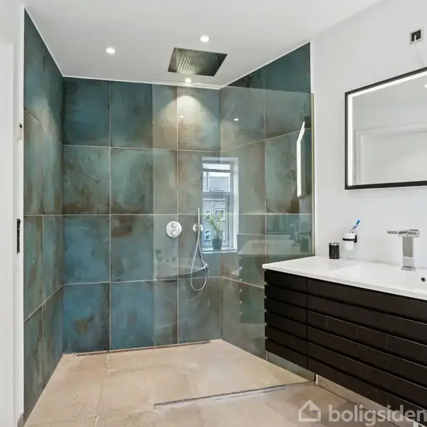 Bruseniche med stor kvadratisk brusehoved og glasvæg. Ved siden af en sort skabsenhed med dobbeltvask og spejl. Rummet har moderne fliser og spotlys i loftet.