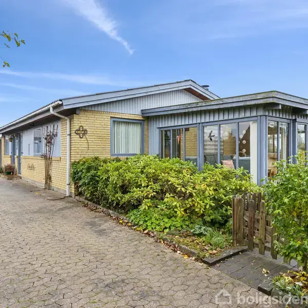 Et enfamiliehus med gul murstensfacade og store vinduer står omgivet af grønne buske og en flisebelagt indkørsel. Blå himmel og træer i baggrunden giver en fredelig atmosfære.