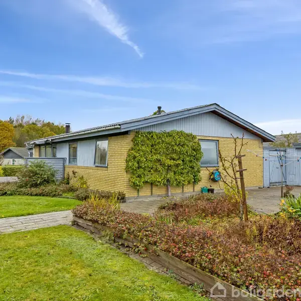 Et gulstenshus med fladt tag står på en velplejet græsplæne omgivet af buske og træer under en blå himmel.