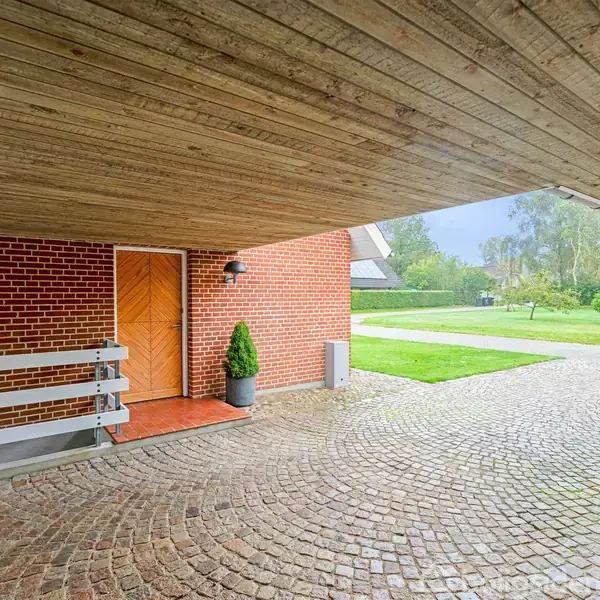 Carport med røde murstensvægge og træloft, der beskytter en indgangsdør med en lanterne. Foran ses en brolagt indkørsel, der fører til en grøn græsplæne med træer i baggrunden.