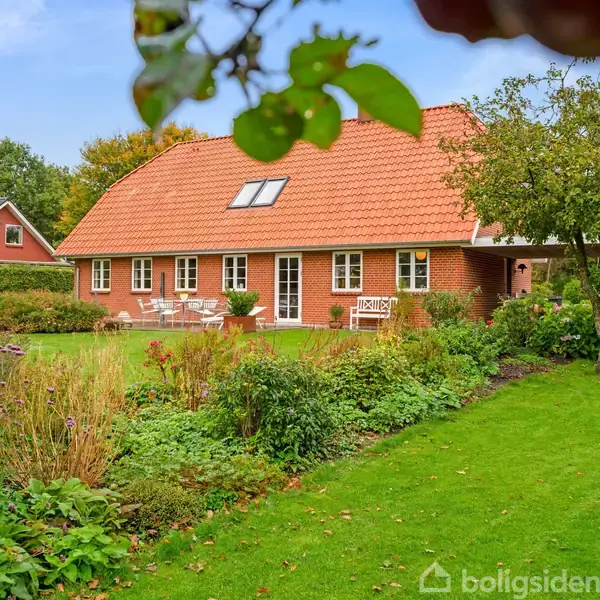 Et rødt murstenshus med rødt tegltag står omgivet af frodige buske og en grøn græsplæne i en have med blomsterbed og havemøbler.