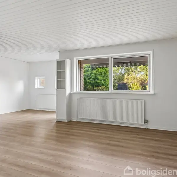 Stue med lyst trægulv uden møbler, store vinduer der slipper naturligt lys ind, hvid loft og vægge, en åben reol ved vinduet og en radiator under, omgivet af grønt udendørs.