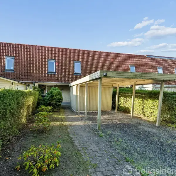 Carport står foran et rækkehus med rødt tag, omgivet af hække. Gårdspladsen har fliser og grus, og himlen er klar med spredte skyer.