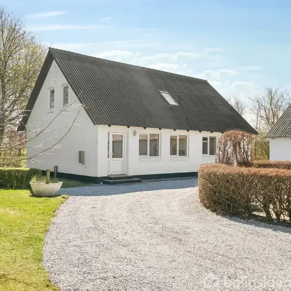 Et hvidt hus med sort tag står stille omgivet af en grusvej og grønne træer i en have. En busk skaber en naturlig indgang langs vejen.