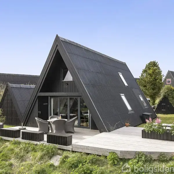 Et sort A-formet hus med store vinduer står på en træterrasse med havemøbler. Omgivet af grønne planter og græsplæner under en klar blå himmel.