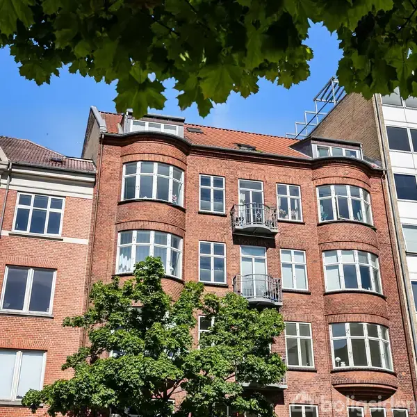 Murstensbygning med buede vinduer og en lille balkon. Træ med grønne blade ses foran bygningen. Andre bygninger omgiver den i en bymiljø. Øverst ses teksten "Anno 1910".