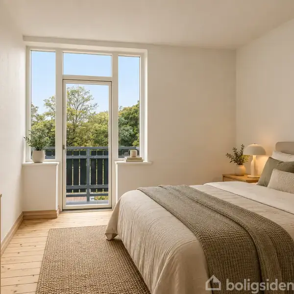 Soveværelse med en dobbeltseng dækket med grå puder og tæppe, ved siden af et natbord med lampe. Store vinduer åbner ud mod en balkon med udsigt til træer.