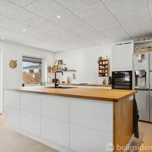 Et moderne køkken med en stor, hvid køkkenø fyldt med skuffer, en indbygget ovn, stål-køleskab og åbne hylder. Udenfor ses en snedækket have gennem store vinduer.