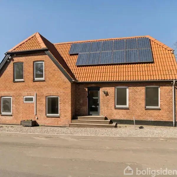 Rødt murstenshus med solpaneler installeret på det orange tegltag, står på en asfalteret vej. Vinduer er jævnt fordelt på facaden; en hvid stakit indhegner baggrunden.
