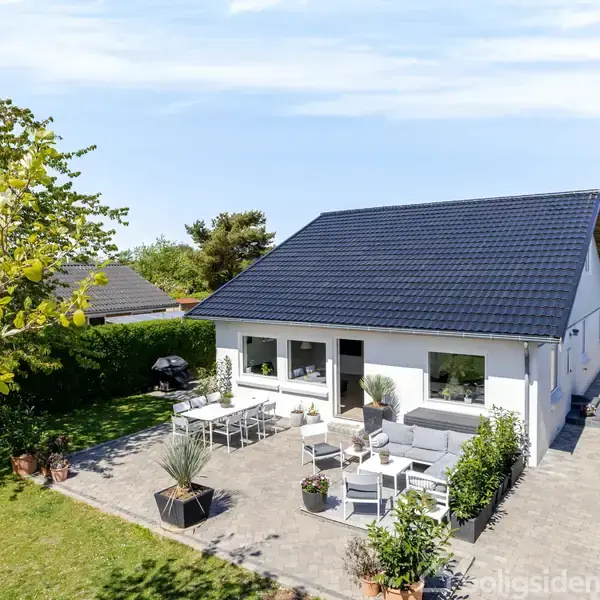 Et hvidmalet hus med mørkt tag står i en frodig have. En stor terrasse foran huset har havemøbler. Omgivet af grønne buske og træer under en klar blå himmel.