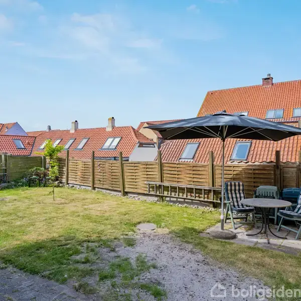 En terrasse med parasol og havemøbler står i en have med græsplæne, omgivet af et hegn. Bagved ses rækkehuse med røde tage under en blå himmel.