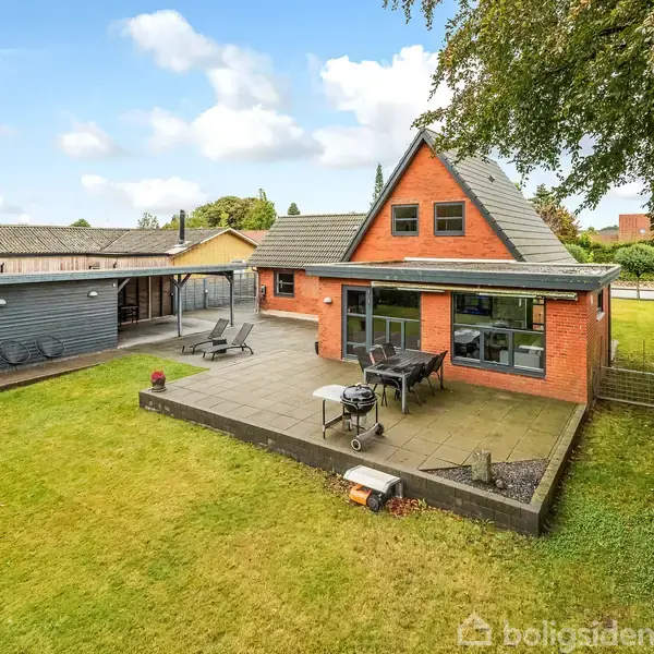 Et rødt murstenshus med store vinduer står i en grøn have. En stor terrasse foran huset har havemøbler og grill. Træer og hegn omgiver grunden.