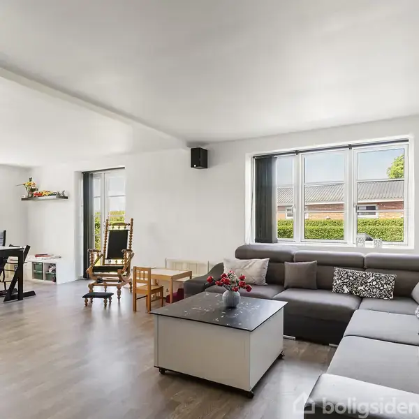 En stue med en stor grå hjørnesofa står under to vinduer. I hjørnet nær vinduerne er der en sort lænestol og et lille bord. I baggrunden er et spisebord og stole med et vindue ud til haven.