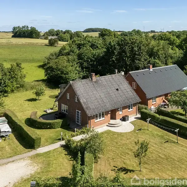Rødt murstenshus omgivet af frodig have og træer. Yderligere bygninger ses nær hovedhuset, der er placeret i et landligt område med marker i baggrunden og en lille sti.