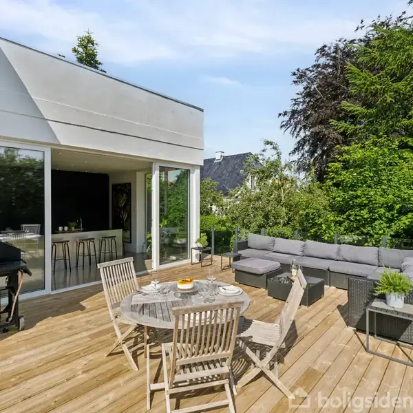 Terrassen har en rund spisebord med træstole, sort grill og en grå hjørnesofa. Den er omgivet af træer og buske, og store vinduer fører ind til huset.